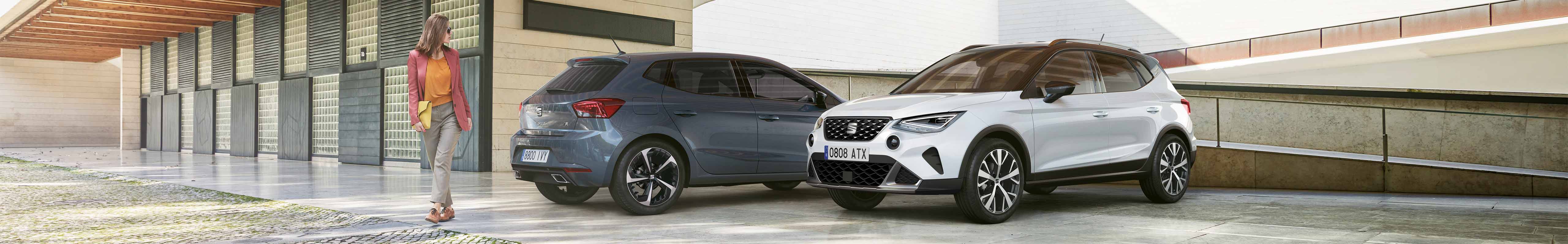 Woman walking between a SEAT Ibiza in blue, magnetic tech and a SEAT Arona in Nevada White and Midnight Black, after completing OTA updates